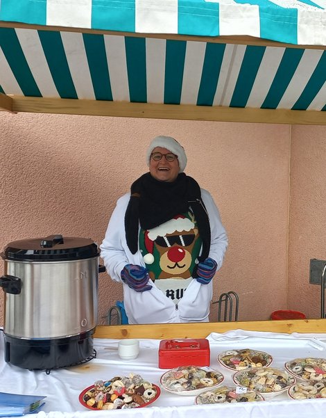 Mitarbeiterin mit Weihnachtspulli und Zipfelmütze verkauft Kekse und Punsch