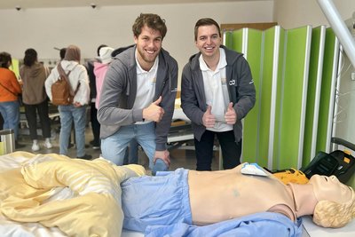 Zwei Auszubildende mit Daumen hoch am Krankenbett mit Pflegesimulator