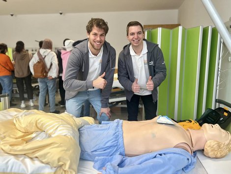 Zwei Auszubildende mit Daumen hoch am Krankenbett mit Pflegesimulator